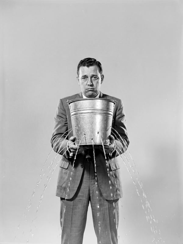 A resigned businessman holding a metal bucket that sprays water from every side. A black-and-white picture in a 1960s studio style.