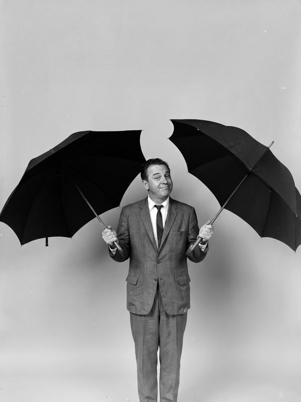 A pleased man indoors holding two open umbrellas at once. A black-and-white picture in a 1960s studio style.